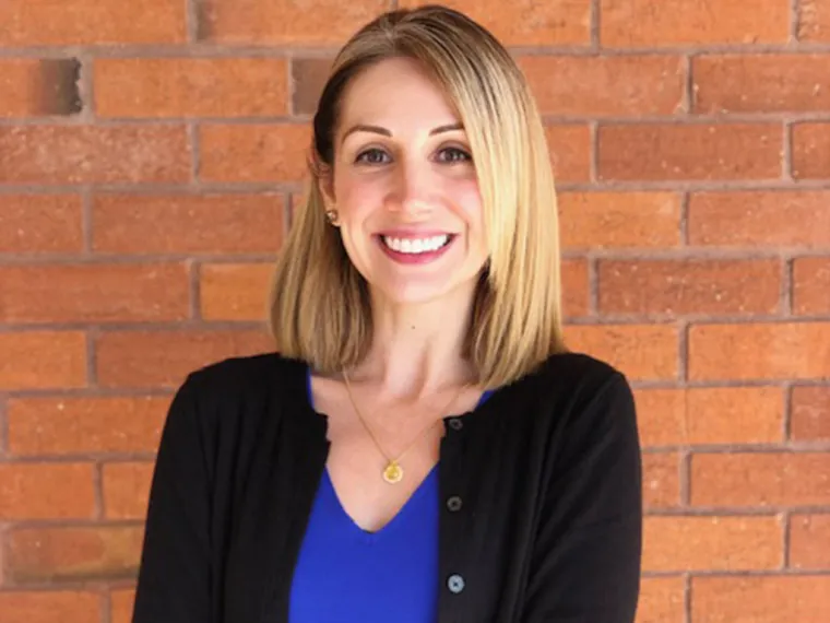 Meghan McKenney smiling for photo, wearing black shoulder cover and blue blouse, brick wall in background