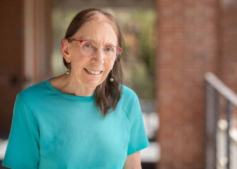 Ellen Fishman smiling for photo, wearing teal blouse and earnings, and red rimmed glasses